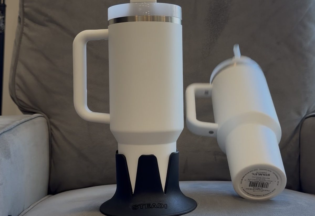 A 40 oz tumbler standing upright in a black Steadi on a grey armchair cushion; the bare tumbler behind it has tipped onto its side.