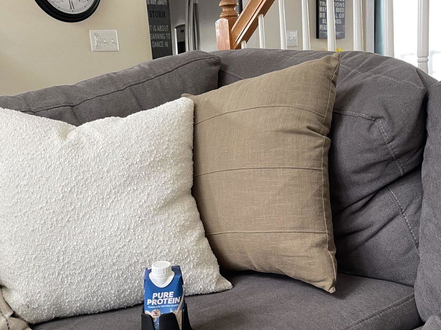 A black Steadi holding a protein shake on the seat cushion of a grey sectional between pillows.