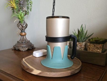 A gold tumbler with a striped straw in a turquoise Steadi on a round wooden board beside a lamp.