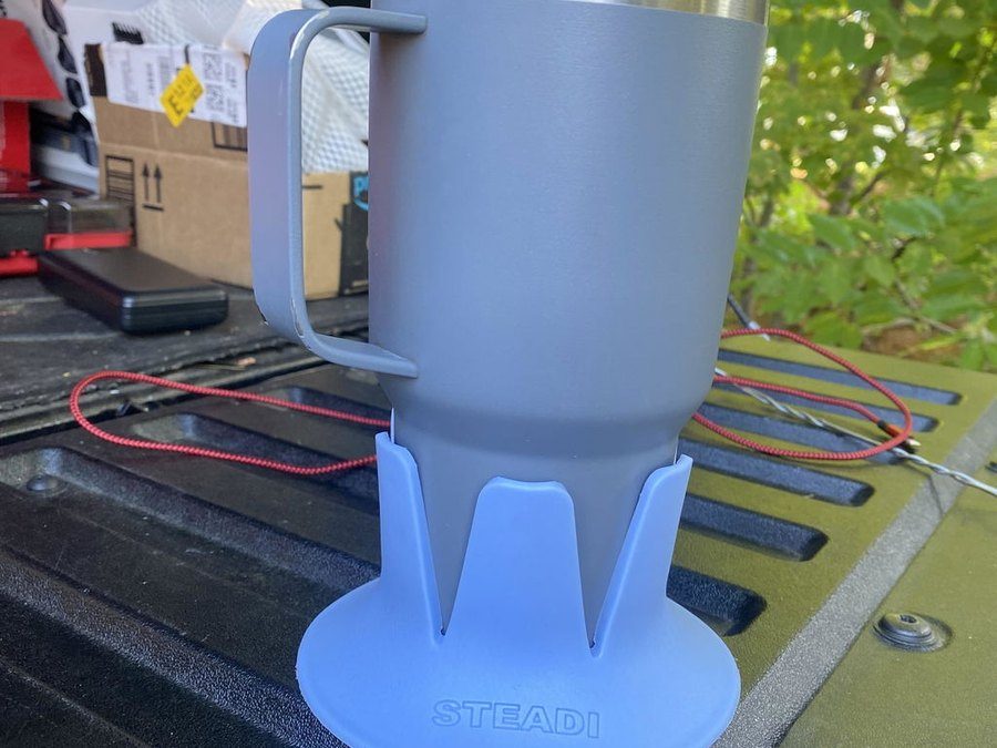 A grey handled tumbler in a periwinkle Steadi on a pickup bed rail, trees behind.