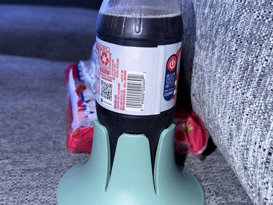 A plastic cola bottle in a mint Steadi on a grey woven couch cushion beside the arm.